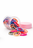 A Pret my baby Pack of 100 everyday mix hair ties for medium hair, in pink, orange, blue, and gray, spills from a clear jar with a pink lid onto a white surface.