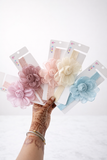 A hand with mehndi holds five Pret my baby Fascinator extra large flower with rose bud baby headbands in pastel pink, cream, and blue on white packaging. The soft background highlights these adorable infant accessories.