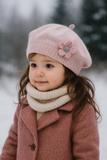 A young girl wears the Pret my baby Soft Woollen knitted Beret cap with flower, paired with a cream scarf and pink coat, standing outdoors in snow with blurred trees—a perfect winter look for toddlers.
