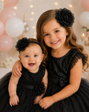 Two young girls smile together in matching black dresses and pretmybaby’s Flower bloom fascinator sibling set. The older sister hugs her baby sibling, pastel balloons softly blurred in the background.