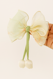 A hand holds the Pret my baby Medium fascinator tulip bow, featuring pastel organza, soft green ribbon tails, and two white fabric cherries. The plain light background highlights its gentle spring-inspired charm.