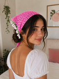 A young woman with dark hair in a loose bun wears the pretmebabe Premium crochet bandana scarf band in bright pink, adorned with small white daisies, styled as a beach hair band for sun protection. She pairs it with a puff-sleeve top and gold hoop earrings, smiling indoors.
