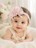 A happy baby in a cream outfit wears the Pret my baby Fancy fascinator vintage bow and pearls headband, featuring lace and chiffon flowers, while lying on a soft blanket with blurred flowers in the background.