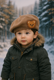 A young child in a green winter coat wears the Pret my baby Soft Woollen Knitted Beret cap with a teddy bear design, standing outdoors as snow gently falls in a snowy forest.