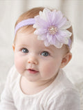 A baby with light hair and blue eyes wears the Pret my baby Fascinator large ombre flower baby headband in lavender, featuring a big organza flower. Dressed in a pale shirt, the baby gazes slightly upward with a gentle expression.