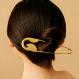 A person with dark hair in a low bun, secured by the Pretmebabe Safety Pin bun clip, a large gold metal hair accessory, shown against a beige background.