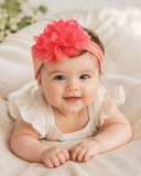 A smiling baby in a cream outfit wears the Pret my baby Soft cotton everyday baby headband with a large flower, lying on a light blanket and looking up with bright eyes—the perfect accessory for a newborn photoshoot.