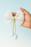 A hand holds the Pret my baby Medium fascinator tulip bow—light blue organza with subtle sparkle, green ribbon tails, and tulip-like flower accents—set against a soft blue background.