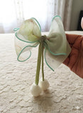 A hand displays the Pret my baby Medium fascinator tulip bow—a delicate, translucent green organza accessory with floral patterns and green edging, featuring two small white tulip-like buds on green stems, set against a lace-covered table.