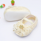 Pret my baby’s Rosette bellies with star in cream feature gold glittery stars and a front bow. One shoe is upright, the other flipped to display its white sole. Small potted plants in the background add charm—perfect for gifting!.