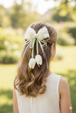 A girl with wavy brown hair wears a white dress and the "Tulip with pearls and delicate ribbon fascinator hairclip" by Pret my baby, featuring artificial tulips, pearls, and ribbons, while standing outdoors in a sunlit garden.